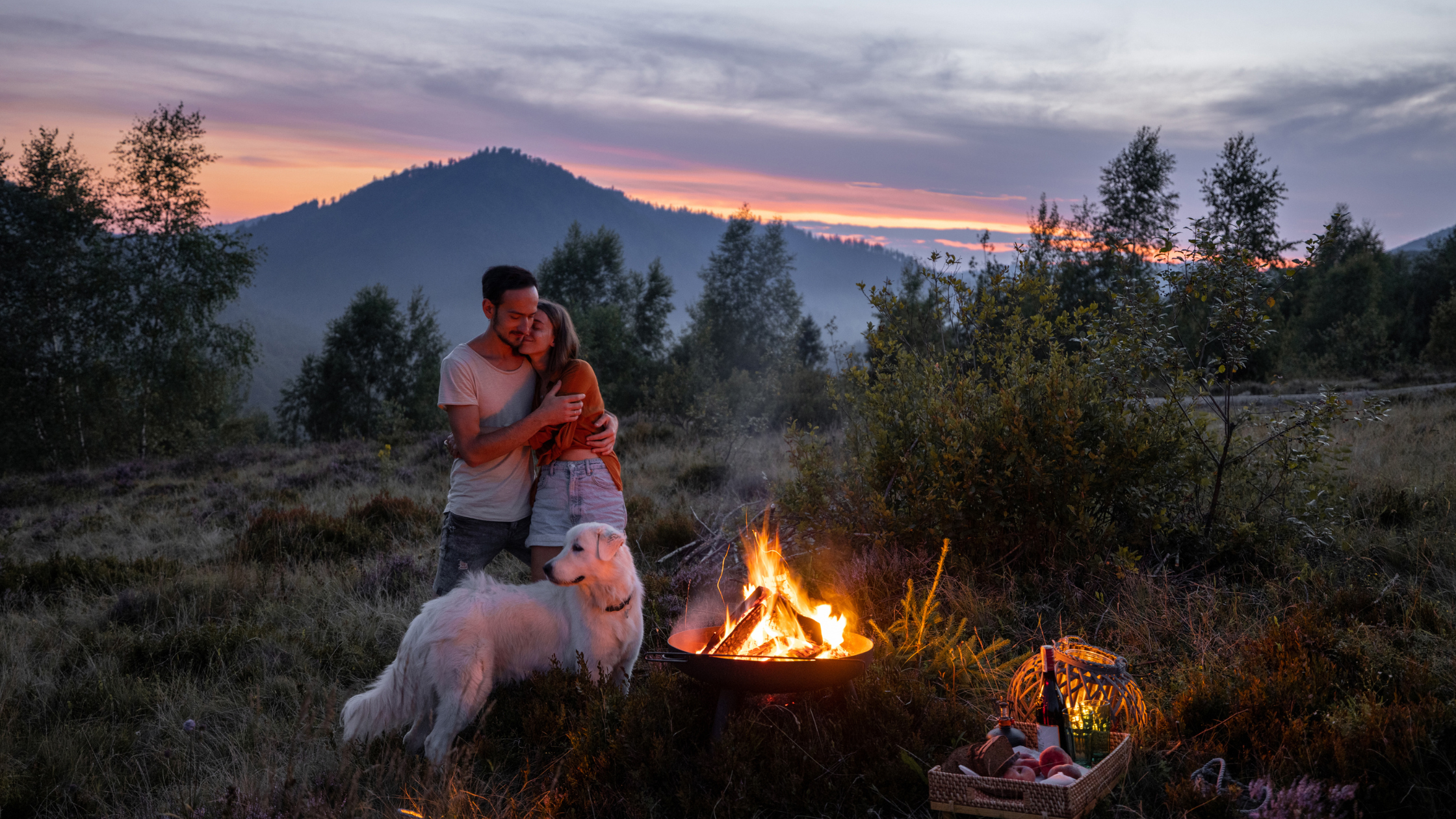 Ein Päärchen steht umschlungen mit ihrem Hund am Lagerfeuer, im Hintergrund Berge